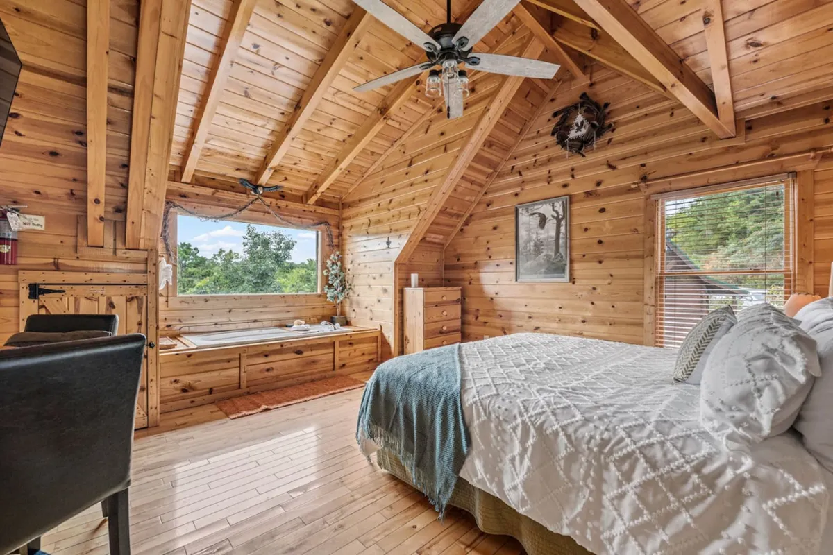 A tranquil bedroom space with a wooden ceiling, a large window framing serene outdoor views, a bed with plush bedding, and built-in bench seating. The mood is peaceful, offering a restful atmosphere ideal for individuals or couples seeking quiet and comfort.