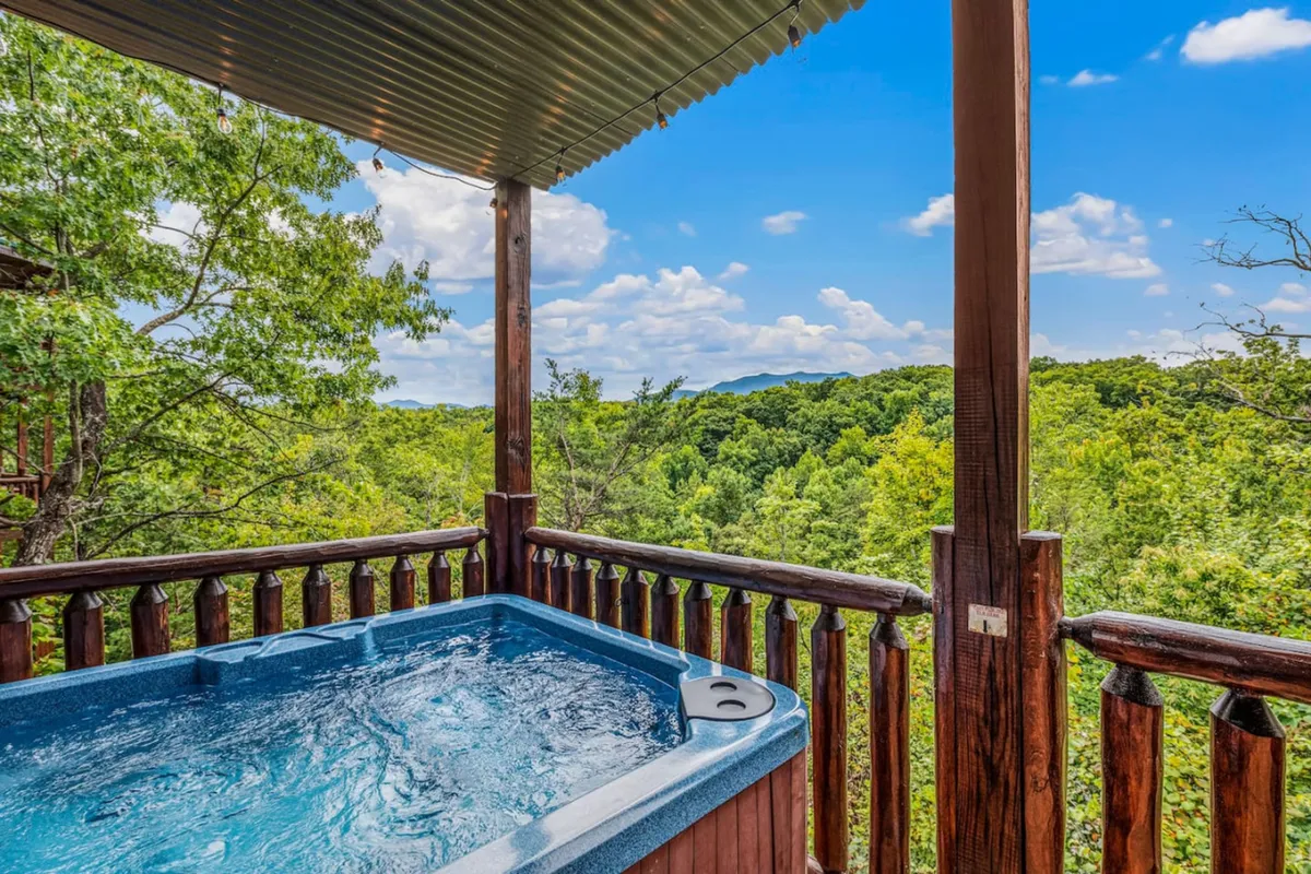 This image showcases a bubbling hot tub set on a wooden deck, framed by a sturdy railing. In the background, lush greenery and distant mountains are visible, creating a serene outdoor setting ideal for relaxation or socializing. The tranquil atmosphere is enhanced by the natural light and peaceful surroundings.