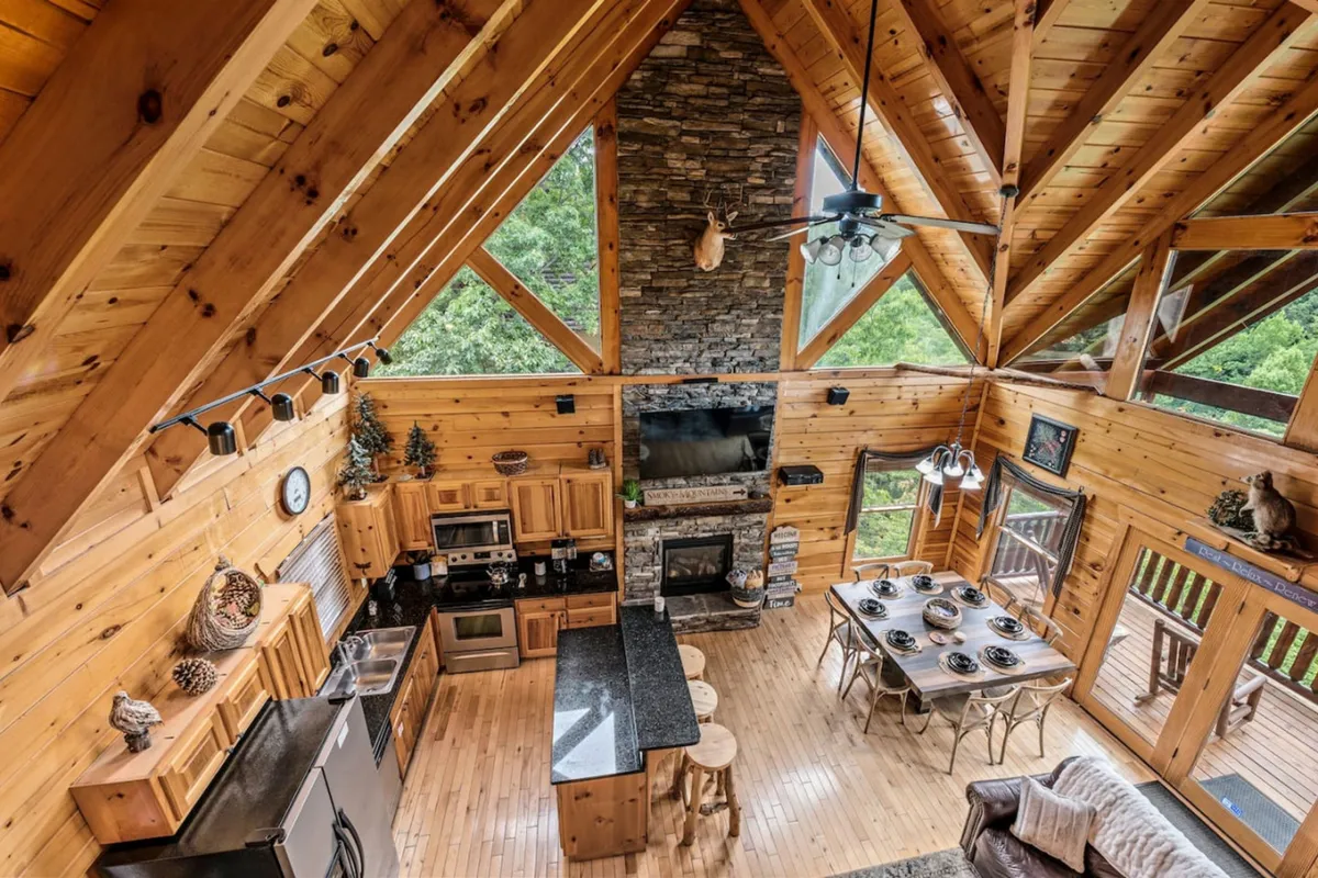 A central view of the open-concept kitchen, dining, and living area. The expansive windows flood the space with natural light and frame beautiful mountain views. The stone fireplace anchors the cozy seating and dining area, while the kitchen island and dining table invite gathering for meals or conversation.