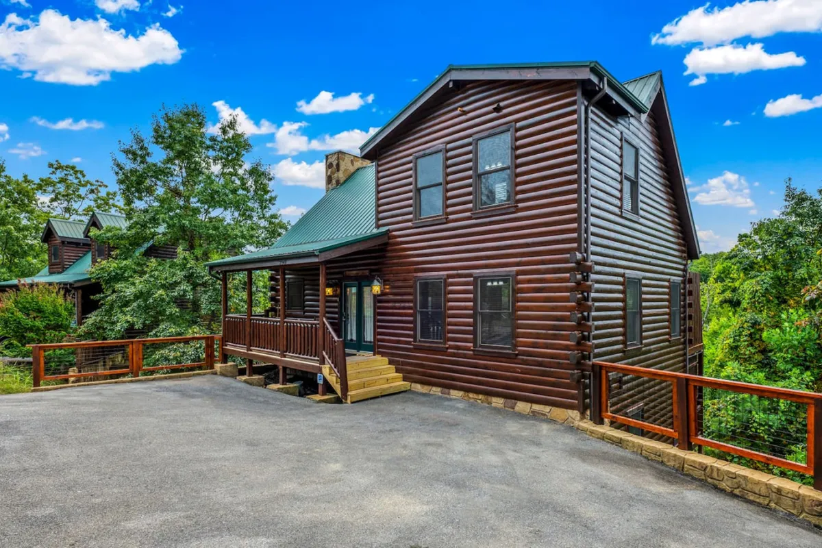 An inviting log cabin with a green roof features a spacious wooden deck surrounded by trees. The deck provides plenty of room for gatherings, barbecues, or alfresco dining. The natural setting and cozy structure suggest an ideal place for outdoor living and lively get-togethers.