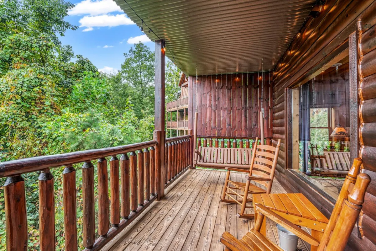 This exterior image features a rustic log cabin porch with wooden rocking chairs, a sturdy railing, and lush greenery surrounding the cabin. The serene setting is framed by the natural beauty of the Smokies, suggesting privacy, peace, and a connection to nature. The porch invites moments of stargazing, quiet conversations, or sharing a morning coffee while immersed in the woods.