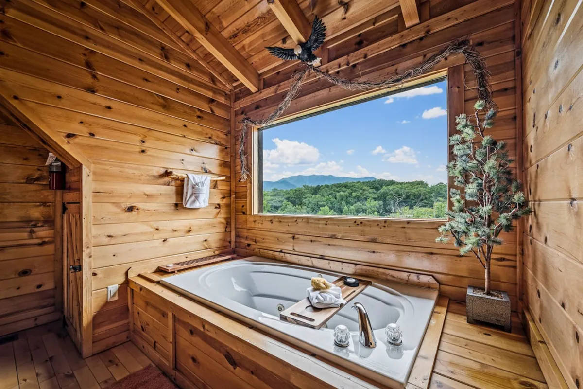 A serene interior shot of a spacious bedroom featuring a large window with panoramic mountain views, a luxurious jacuzzi tub set within the bedroom, warm wooden wall paneling, and a decorative plant that adds a touch of nature. The mood is tranquil, and the in-room tub is a central feature, inviting relaxation while taking in the landscape.