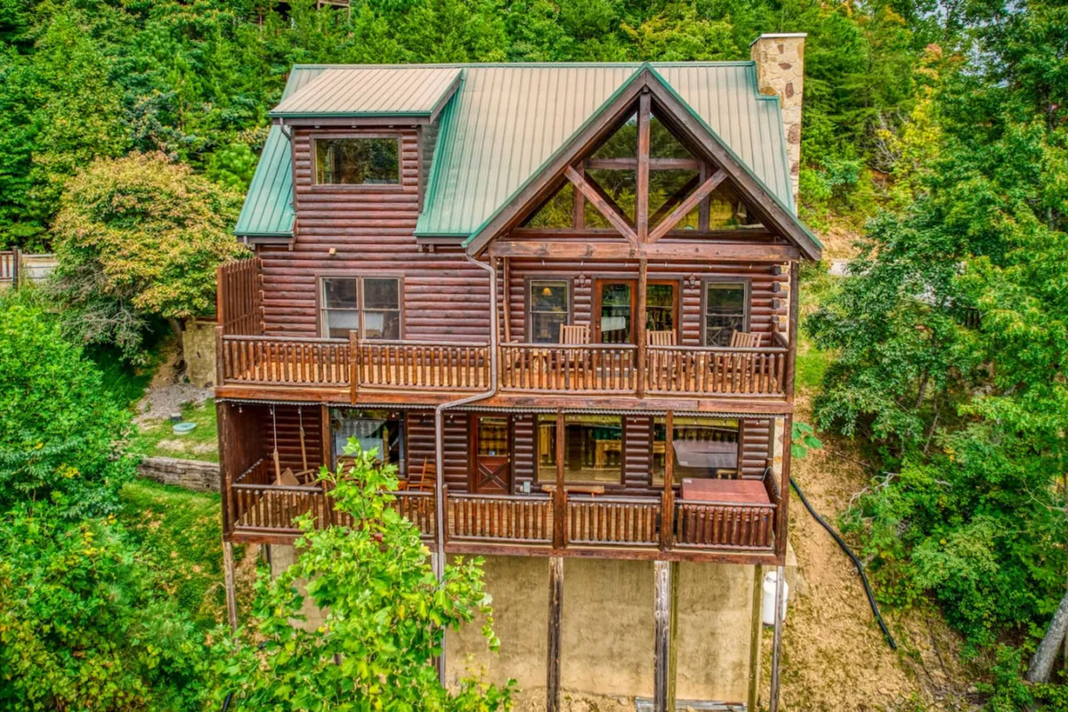 A wide shot of the log cabin exterior with a green metal roof, featuring wooden balconies and surrounded by lush trees. The multi-level design is visible, emphasizing the spaciousness and privacy offered by the layout.