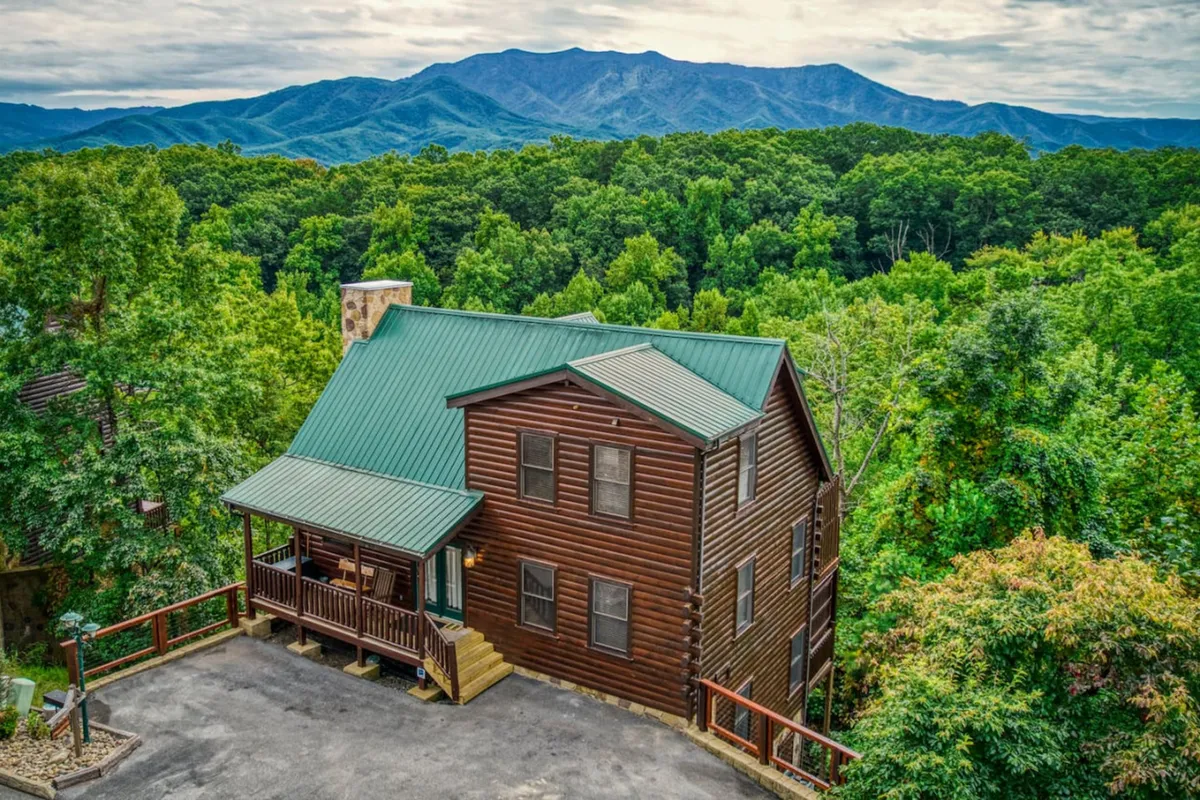 The hero image features a charming log cabin with a green metal roof, nestled among lush trees and surrounded by rolling Smoky Mountain views. The inviting front porch hints at sunrise coffees, while the dramatic natural setting evokes peace and adventure.
