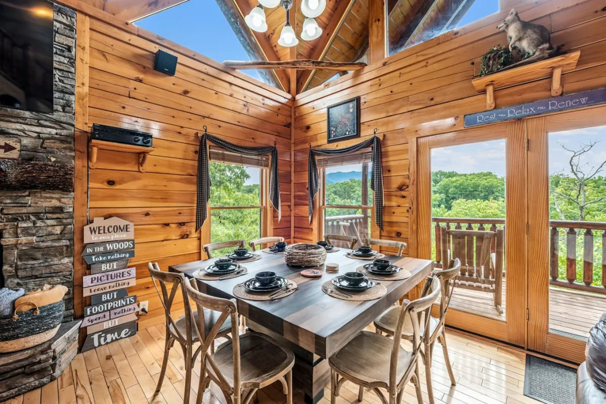 A welcoming dining area with a table set for a meal, bathed in natural light from large windows. Wooden beams and a stone fireplace add warmth, and the view through the windows hints at the surrounding mountain scenery.
