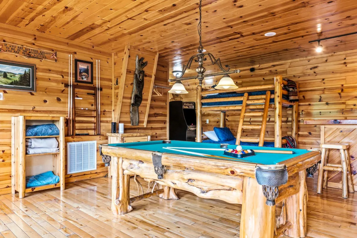 A spacious interior with a central pool table, wooden shelves lining the wall, a stylish chandelier overhead, and a comfortable lounge area. The mood is playful and relaxed, inviting guests to gather and enjoy games or simply socialize in comfort.