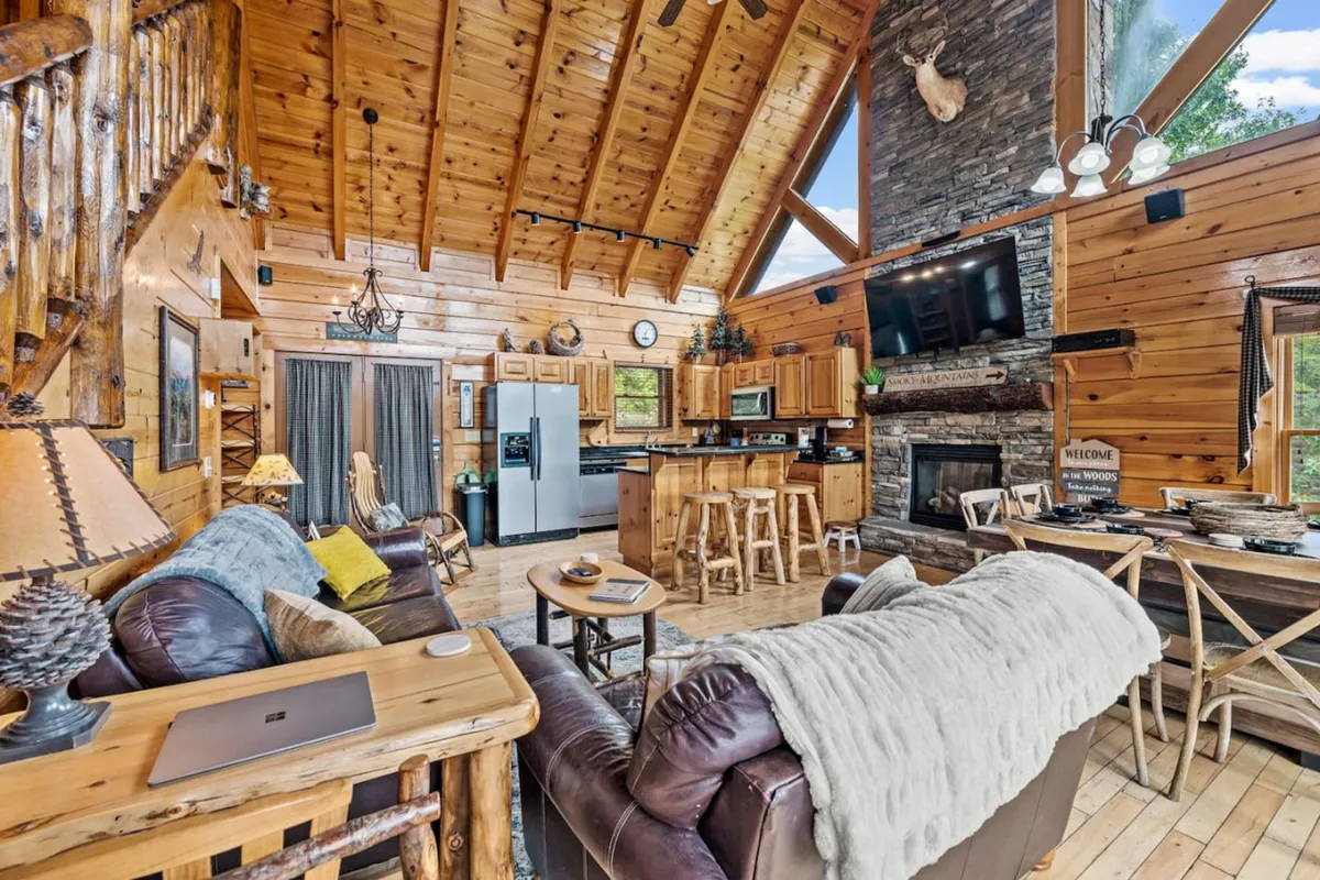 A wide-angle shot of the living and kitchen area, featuring a grand stone fireplace, exposed wooden beams, a kitchen island, and a comfortable seating area. Light streams in through large windows, and modern amenities are subtly visible.