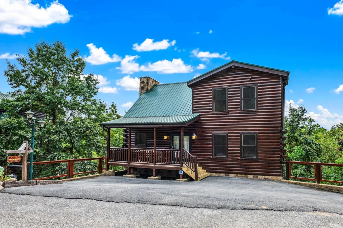 A classic log cabin structure with a striking green metal roof and a welcoming wooden porch, surrounded by lush trees. The porch features comfortable seating and offers views of the natural landscape, hinting at tranquil mornings and relaxing evenings.