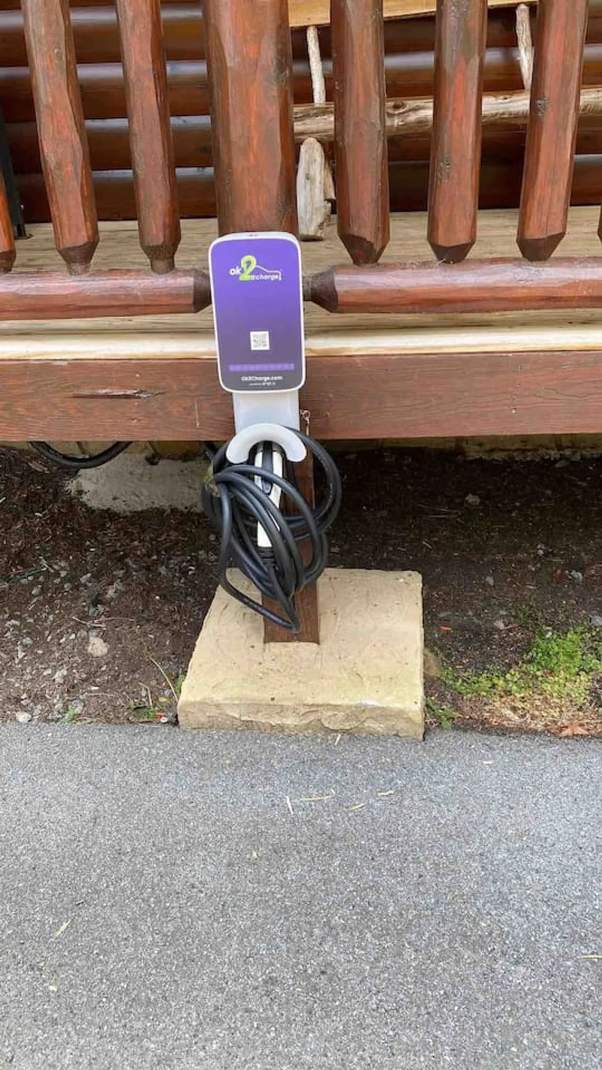 An exterior shot featuring a dedicated electric vehicle (EV) charger set against a rustic wooden railing with a concrete base. The outdoor setting emphasizes eco-friendly travel and the integration of modern technology with the cabin's natural environment.