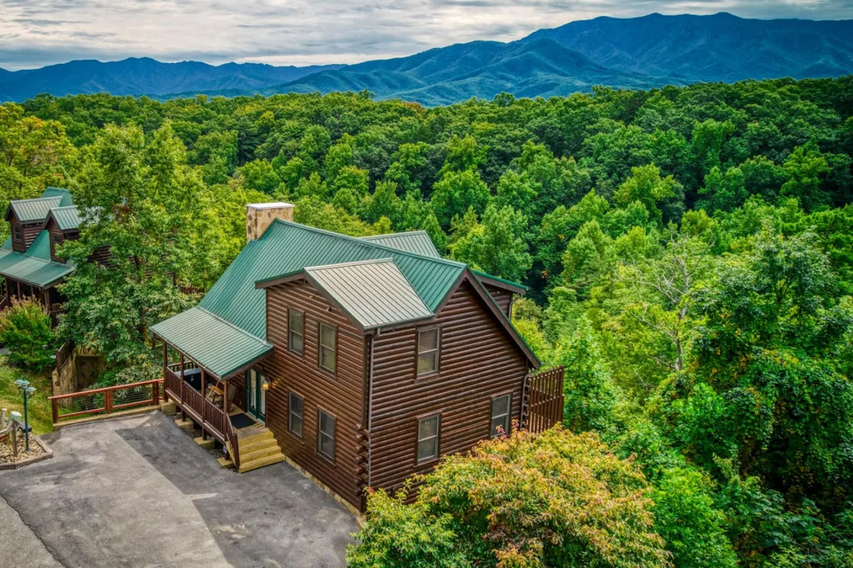 A classic log cabin with a green metal roof is nestled among tall trees, with the dramatic silhouette of the Smoky Mountains creating a stunning natural backdrop. The property’s rustic charm and forested setting evoke a sense of escape and adventure, perfect for nature lovers.