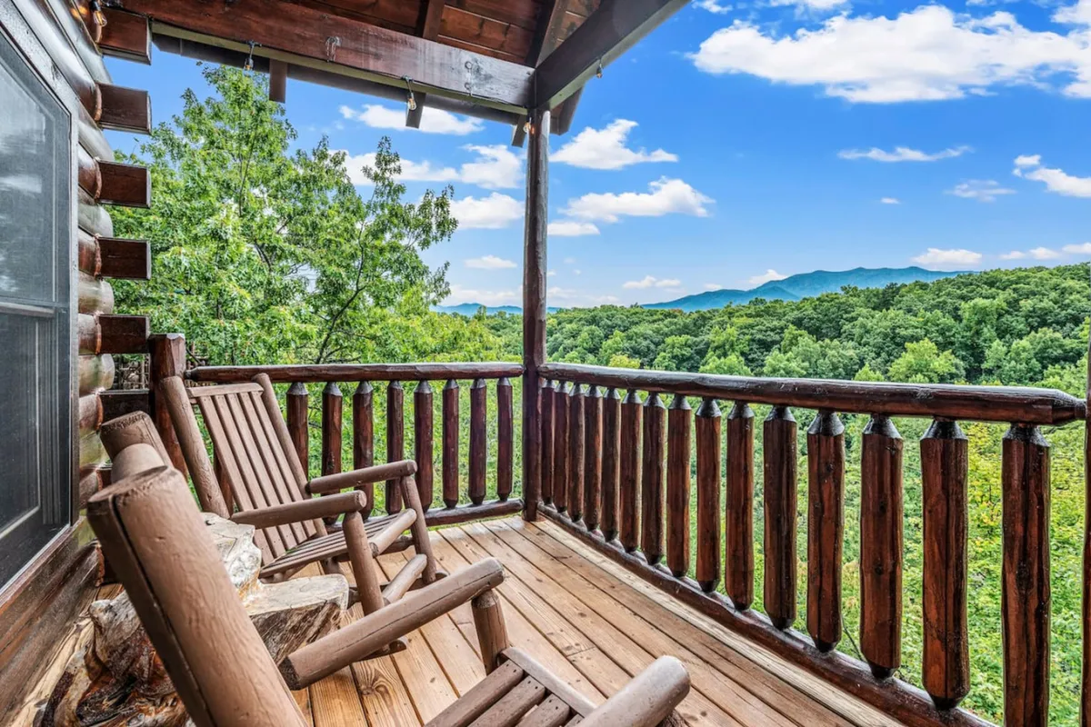 A pair of wooden rocking chairs sit on a deck with a wooden railing, looking out toward distant mountains and lush green treetops. The space exudes tranquility and is perfect for enjoying quiet moments, sunrises, or casual conversations while being immersed in nature.