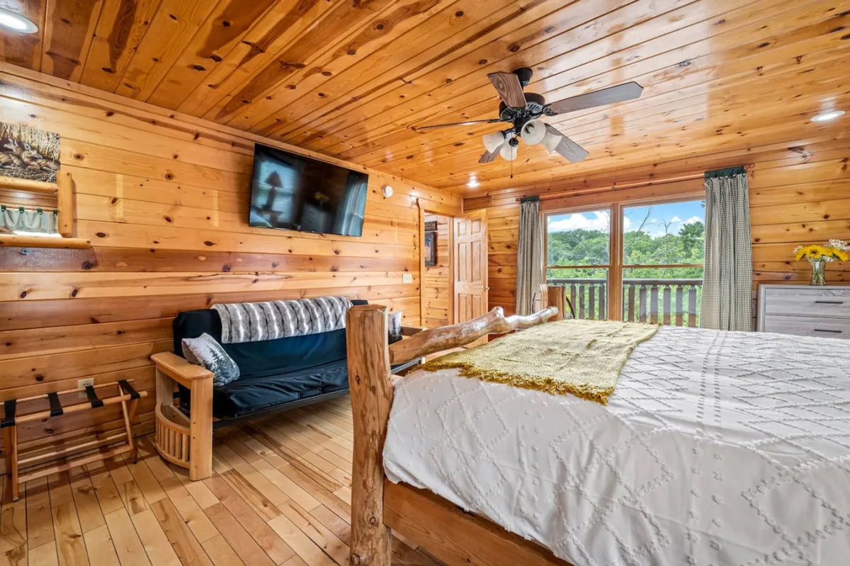 A cozy, inviting bedroom with wooden walls, a bed adorned with a decorative blanket, a comfortable sofa with throw pillows, a TV mounted on the wall, and a large window offering scenic views. The space blends comfort and functionality, supporting family or group stays.