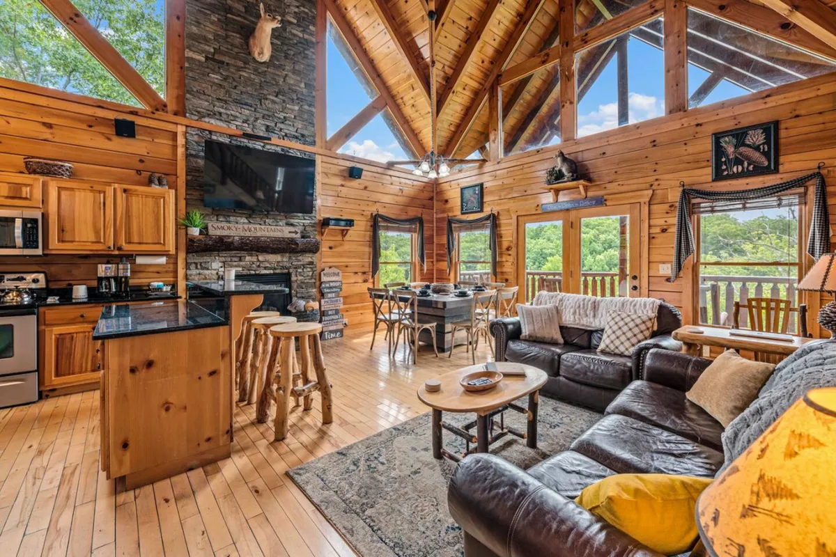 A welcoming interior with exposed wooden beams, a majestic stone fireplace, expansive windows, and a cozy seating area. The large windows offer glimpses of the natural surroundings, blending the comfort of the inside with views of the outdoors.